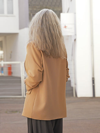 Person von hinten im urbanen Umfeld, trägt den OFF#DLY Blazer Dana camel. Der Blazer hat eine moderne Silhouette mit Reverskragen und aufgesetzten Taschen, kombiniert mit dunkler Hose.