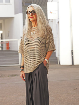 Ältere Frau in lockerem La Haine Tee 1226 mud mit Netzstruktur und Oversize-Schnitt, kombiniert mit weitem, dunklem Beinkleid, steht in modernem, architektonischem Umfeld und trägt stilvollen Schmuck.