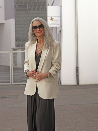 Ältere Frau trägt den OFF#DLY Blazer Dana beige in einer urbanen Umgebung. Der leicht übergroße Blazer hat aufgesetzte Taschen und einen klassischen Reverskragen, stilvoll kombiniert mit Schmuck.