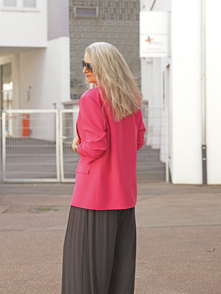 Eine reife Frau trägt den OFF#DLY Blazer Dana pink mit klassischem Reverskragen und aufgesetzten Taschen in urbaner Umgebung, kombiniert mit schwarzen Hosen und Sonnenbrille.