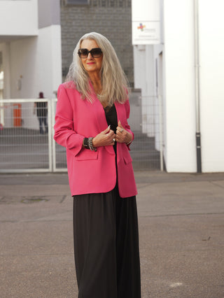 Mature Frau im urbanen Umfeld trägt den OFF#DLY Blazer Dana pink, ein zeitloses Essential mit klarer Silhouette, Reverskragen und aufgesetzten Taschen, über schwarzem Top und weiter Hose.