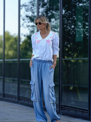 Frau trägt Gitta Banko Tencel Pant Emily Nevada Sun, lockere hellblaue Cargo-Hose mit großen Seitentaschen, kombiniert mit weißer Bluse, in urbaner Umgebung.