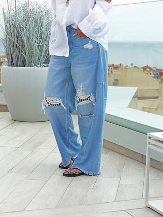 Person trägt OFF#DLY Denim Ibiza lightblue: Weite, hellblaue Jeans mit großen, weißen Spitzeneinsätzen an den Knien und fransigem Saum, kombiniert mit schwarzen Sandalen auf einer Holzterrasse.