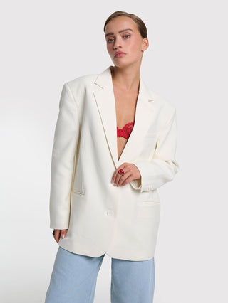 Eine junge Frau trägt den ALIX Flowy Oversized Blazer cream, offen über einem roten Spitzen-Bralette, kombiniert mit hellblauen Jeans. Ein eleganter, moderner Look mit klarem, hochwertigem Design.