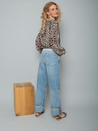Emily Bluse 157500 leo: Junge Frau trägt eine langärmelige, locker sitzende Shirtbluse mit Leo-Print und elastischem Strickbund, kombiniert mit weiten Jeans und flachen Sandalen, im Studio fotografiert.