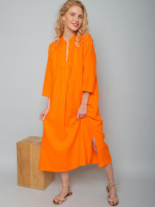 Emily Kleid 156770 orange zeigt eine Frau in einem leuchtend orangefarbenen, locker geschnittenen Leinenkleid mit V-Ausschnitt, dreiviertel Ärmeln und seitlichem Schlitz, getragen mit hellen Sandalen.