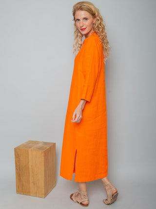 Frau trägt das Emily Kleid 156770 orange von Emily van den Bergh, ein langes, locker geschnittenes Leinenkleid mit V-Ausschnitt, Faltendetails und Seitenschlitzen, kombiniert mit beige Sandalen.