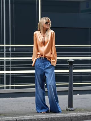 Junge Frau in weiter Vivi Denim Gold Rush Jeans von Gitta Banko, kombiniert mit einer oversized Bluse, auf modernem Gehweg stehend, urbanes Mode-Statement.