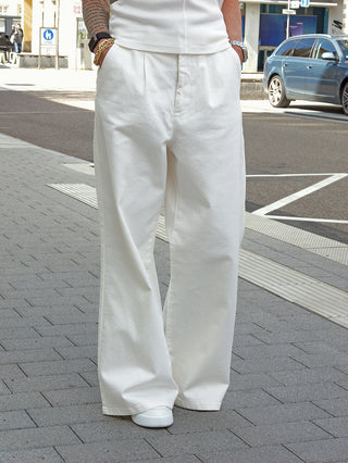 Person trägt die Gitta Banko Hose Rylee warm white mit weitem, fließendem Schnitt. Die weiße Denim überdeckt weiße Canvas-Sneaker. Hände in Taschen, Armbänder sichtbar. Elegante, lässige Silhouette.