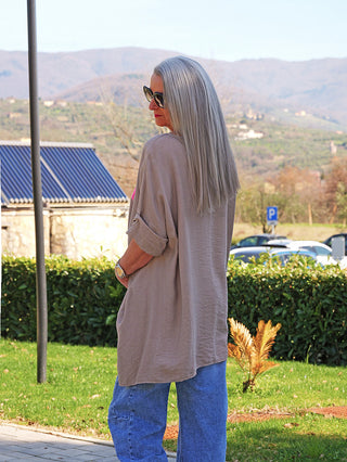 Frau im Profil trägt OFF#DLY Longshirt Tamara taupe, locker geschnitten mit krempelbaren Ärmeln, stehend auf einem Gehweg neben Hecke, vermittelt lässigen, sportlichen Look in ländlicher Umgebung.