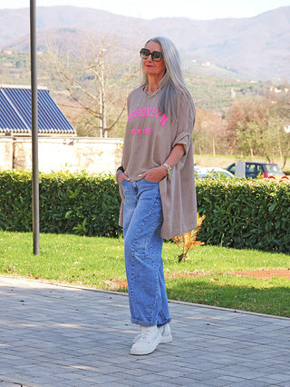Frau trägt OFF#DLY Longshirt Tamara taupe mit „BROOKLYN NEW YORK“-Print in Neonpink, kombiniert mit blauen Jeans und weißen Sneakers, während sie auf einem gepflasterten Weg steht.