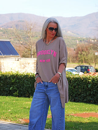 Person im Garten trägt das OFF#DLY Longshirt Tamara taupe mit BROOKLYN in Neon-Pink, kombiniert mit blauen Jeans, silbernem Schmuck und Sonnenbrille; sportlich-lässiger Look.