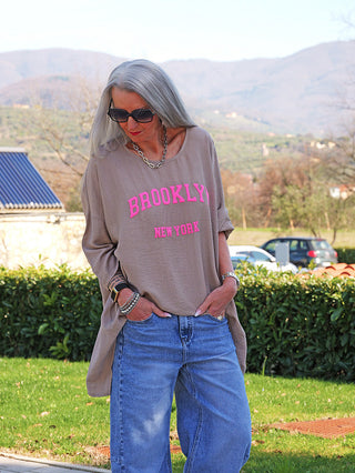 Person in OFF#DLY Longshirt Tamara taupe mit „BROOKLYN NEW YORK“ Print, trägt blaue Jeans. Entspannte Pose mit Händen in Taschen, draußen auf einem Weg neben Hecke.