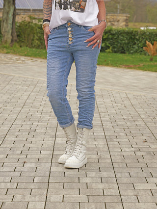 Person trägt OFF#DLY Denim Franka midblue Jeans mit markanter Knopfleiste und aufgerollten Säumen, kombiniert mit cremefarbenen Stiefeln. Sichtbar sind zudem ein T-Shirt, Schmuck und tätowierter Arm.