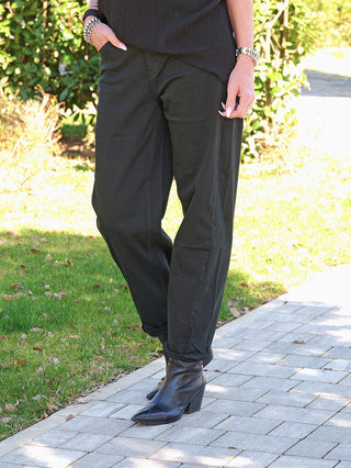 Person trägt OFF#DLY Denim black/black Lola, locker geschnittene schwarze Jeans mit umgeschlagenen Bündchen, kombiniert mit schwarzen Stiefeletten. Eine Hand steckt lässig in der Tasche.