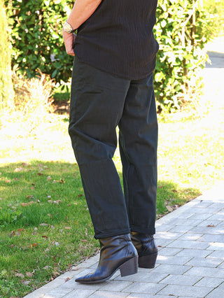 Person trägt OFF#DLY Denim black/black Lola, eine locker sitzende schwarze Jeans mit umgeschlagenem Saum. Kombiniert mit schwarzen Lederstiefeletten, vermittelt sie einen modernen, entspannten Look.