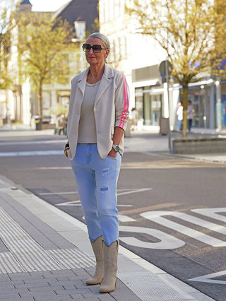 Die Frau trägt den OFF#DLYBlazer Oversized Stripes beige mit pinken Streifen an den Ärmeln, kombiniert mit lässigen Jeans und Stiefeln, was einen modernen, entspannten Streetstyle-Look kreiert.