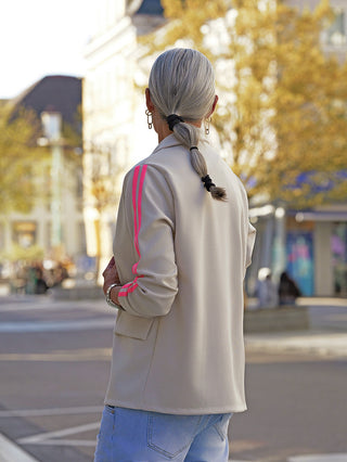 Person von hinten im Stadtzentrum, trägt den OFF#DLYBlazer Oversized Stripes beige mit pinken Streifen an den Ärmeln, kombiniert mit Jeans, zeigt sportlich-eleganten Stil im urbanen Umfeld.