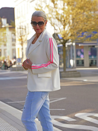Frau in weißem OFF#DLYBlazer Oversized Stripes mit pinken Streifen an den Ärmeln, lässig über weißem Top und Jeans getragen, steht an einer sonnigen Straßenecke.