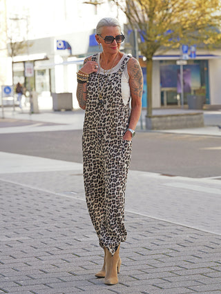 Frau trägt OFF#DLY Musselin Latzhose leo, einen lockeren Leopardenmuster-Jumpsuit über weißem Tanktop, mit tanfarbenen Stiefeln und Accessoires, betont urbanen Stil in lebhafter Einkaufsgegend.