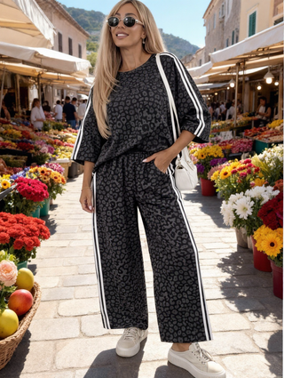 Junge Frau trägt OFF#DLY Anzug Rimini leo/black, bestehend aus lockerem Top und weitem Hose mit weißen Streifen, in einem Blumenmarkt. Kombiniert mit weißen Sneakers und kleiner Schultertasche.