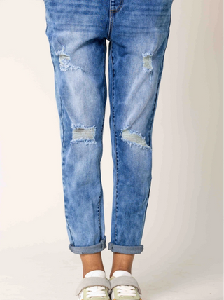 Person trägt OFF#DLY Nina Latzhose midblue mit leicht verwaschenem, zerstörtem Look und gekrempelten Beinen, kombiniert mit weißen und hellgrünen Sneakers mit grauem Riemen.