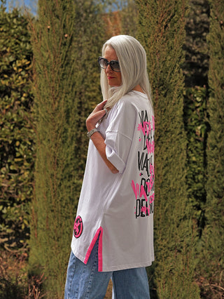 Frau mit langem silbergrauen Haar trägt #VDR T-Shirt 19024 white mit graffitiartigem Rückenprint und pinkem Seitenakzent, kombiniert mit Jeans, zeigt urbanen Stil vor hohen Sträuchern.