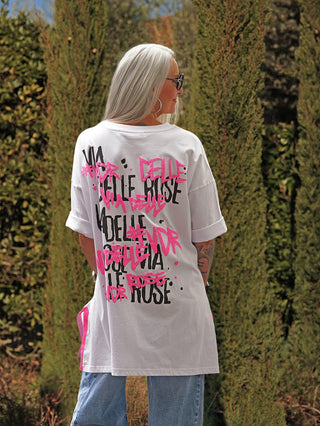 Person mit langem, silbrig-grauem Haar in einem #VDR T-Shirt 19024 white, das einen auffälligen Graffiti-Backprint zeigt, kombiniert mit hellblauen Jeans, steht vor grüner Gartenkulisse.