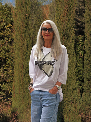 Ältere Person trägt #VDR T-Shirt 19080 white mit Schmetterling und INSPIRE-Print, kombiniert mit blauen Jeans. Accessoires: Sonnenbrille, Creolen, Armbanduhr, Armbänder. Lässiger, moderner Look.