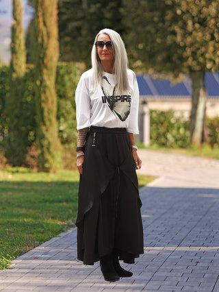 Frau trägt #VDR Rock 19231 schwarz, fließend mit Kordeldetail, das Styling-Variationen ermöglicht. Kombiniert mit weißem Butterfly-Shirt, schwarzen Boots auf Gartenweg, bietet der Rock eine elegante, wandelbare Silhouette.