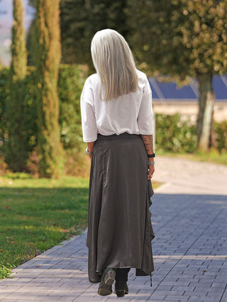 Person von hinten auf einem Gartenweg, trägt den #VDR Skirt 19231 black. Der Rock fällt fließend und wird durch Kordeldetails individuell gestylt, kombiniert mit einer weißen Bluse und Stiefeletten.
