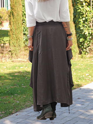 Person trägt #VDR Skirt 19231 black mit asymmetrischem Saum, lockerem Fall und elastischem Bund, kombiniert mit schwarzen Wildleder-Stiefeletten, silbernem Armbanduhr, Armbändern und Tattoos am Unterarm.