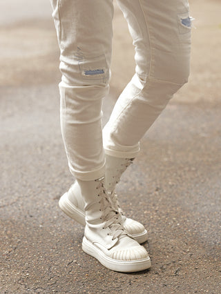 Naheaufnahme von Beinen in schmal geschnittenen, leicht figurbetonten Jeans „OFF#DLY Denim Turin white“ mit dezenten Destroyed-Details und passender heller Schnürstiefel, präsentiert auf einer Gehwegoberfläche.