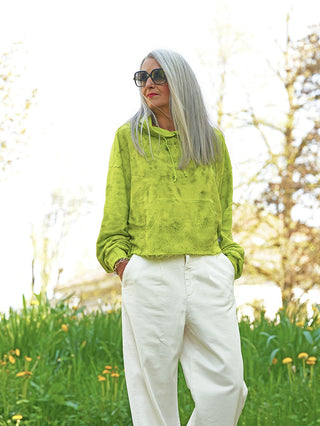 Die Frau trägt den Outryght Celine neon green Hoodie mit Kängurutasche und Kordeln, kombiniert mit weißen, weiten Hosen, in einer Frühlingswiese stehend.