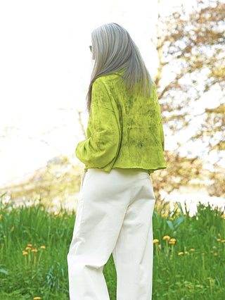 Person von hinten in sonniger Wiese, trägt Outryght Celine neon grünen, lockeren, kurzen Jacke und hochgeschnittene, weiße, weite Hose, umgeben von blühenden Bäumen und gelben Wildblumen.