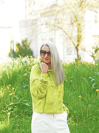 Eine Frau trägt einen Outryght Celine neon green Hoodie, der leuchtend und bauchfrei ist. Sie steht in einer blumenreichen Wiese, mit langen grauen Haaren und großer Sonnenbrille.