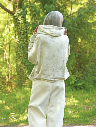 Eine Person im Outryght James Chalk Hoodie steht mit dem Rücken zur Kamera, ihre Hände sanft an der Kapuze haltend, umgeben von einer grünen Waldlandschaft.