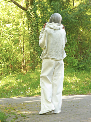 Eine Person trägt den Outryght James chalk Hoodie, leicht oversized, mit weiten weißen Hosen, während sie auf einem sonnendurchfluteten Waldweg steht. Ihre grauen Haare leuchten im Licht.
