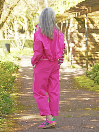 Person trägt OFF#DLY Jacke Eni pink, im lässigen, leicht verkürzten Schnitt, mit großen Brusttaschen und markanter Knopfleiste, auf einem sonnigen Gartenweg stehend, Hände in Hosentaschen.