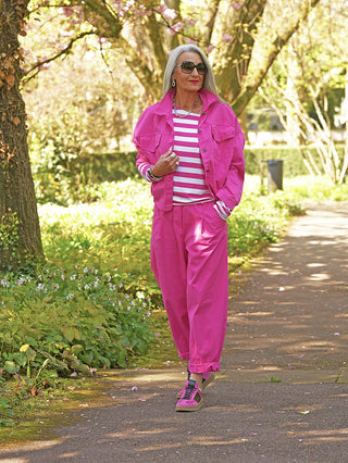 Person trägt die OFF#DLY Jacke Eni pink mit großen Brusttaschen und markanter Knopfleiste, während sie in einem Park spaziert. Die Jacke hat einen lässigen, leicht verkürzten Schnitt.