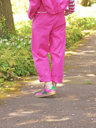 Person trägt OFF#DLY Jogger Sina Pink, eine locker geschnittene Hose mit Bündchen am Knöchel, auf einem Gartenweg. Kombiniert mit passenden Sneakern, umgeben von grüner Vegetation.