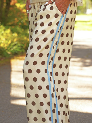 OFF#DLY Pant Dot´s creme/brown zeigt weite, beige Hosen mit großen braunen Punkten und einem schmalen, blauen Seitenstreifen, ergänzt durch eine Hand in der Tasche mit silbernen Ringen.