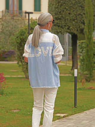 Person von hinten auf Gartenweg, trägt die OFF#DLY Denim Bluse Anna lightblue mit großem LOVE-Schriftzug auf dem Rücken, lockerer Schnitt, transparente Einsätze an den Ärmeln.