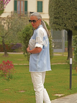 Ältere Frau im Garten trägt die OFF#DLY Denim Bluse Anna lightblue mit Spitzen-Panels, lockerer Schnitt, Knopfleiste, Brusttasche, und „LOVE“-Schriftzug auf der Rückseite, kombiniert mit weißer Hose.