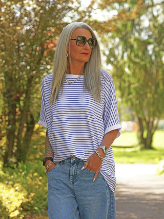 Frau trägt OFF#DLY Ringel Oversized white/lightblue T-Shirt mit schmalen blauen Streifen, entspanntem Schnitt, rundem Ausschnitt und kurzen Ärmeln, kombiniert mit blauen Jeans und Accessoires im Garten.