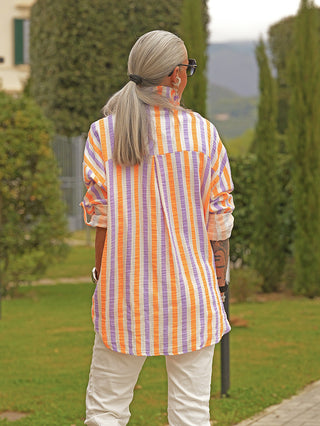 Person von hinten in einem Garten, trägt die OFF#DLY Bluse Dora white/stripe, locker geschnitten mit vertikalen Streifen und Knopfleiste, kombiniert mit off-weißen Hosen.
