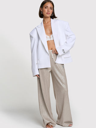 Model trägt ALIX Shiny Pant silver: Weit geschnittene, metallic-glänzende Hose mit elastischem Kordelzug-Bund. Kombiniert mit einem weißen Blazer und Sandalen für einen modernen, sportlich-eleganten Look.