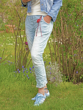 Person trägt OFF#DLY Pant Denim Style Sola lightblue mit leichtem Crinkle-Look. Hose hat hochgekrempelte Bündchen, elastischen Bund und seitliche Taschen, kombiniert mit hellblauen Adidas-Sneakern im grünen Garten.