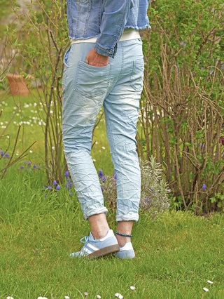 Person trägt OFF#DLY Pant Denim Style Sola lightblue mit schmalem Schnitt und elastischem Bund, kombiniert mit einer Jeansjacke und Adidas-Sneakern, stehend auf einer grünen Wiese.