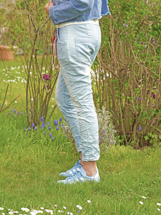Die OFF#DLY Pant Denim Style Sola lightblue zeigt sich als lässige Joggerhose mit Crinkle-Effekt und dekorativen Bändern am Bund, getragen im Garten mit pastellblauen Adidas Sneakern.